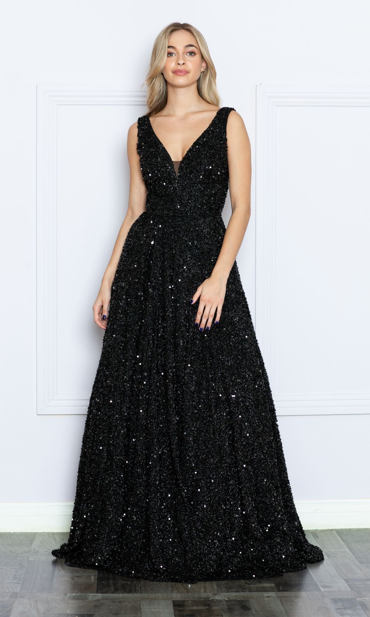 A woman poses against a white wall in the Long Black Sequin A-Line Prom Dress 9168, featuring a sleeveless design, deep V-neckline, and floor-length A-line silhouette for classic evening elegance.